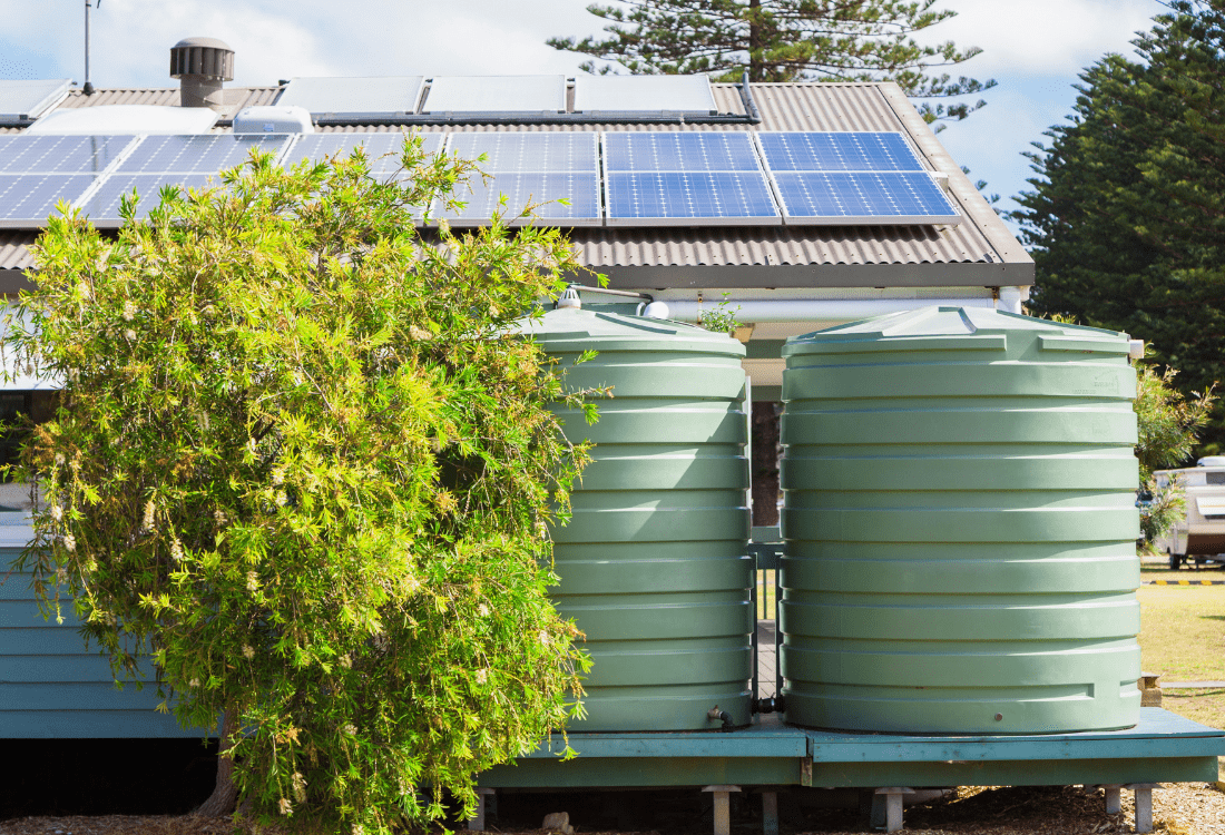 Two green above-ground effluent tanks for wastewater and sludge sit beside a building with solar panels and a bushy shrub in the foreground.